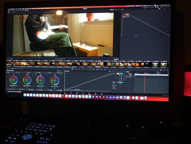 A computer screen displaying a video editing software interface. The software shows various editing tools like color wheels, a timeline with video thumbnails, and graphs. On the screen, a video clip of a person sitting and playing a guitar is visible. The workspace is dimly lit, and a red LED microphone or speaker is positioned on the desk.